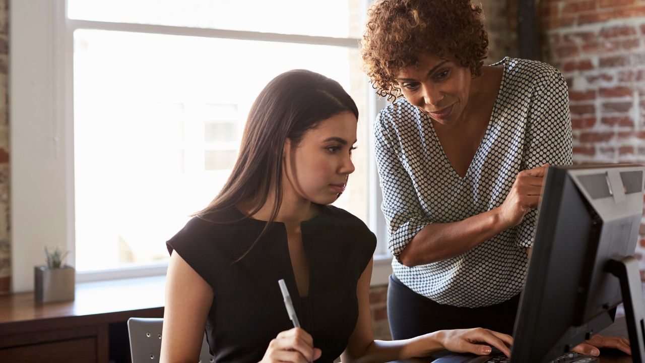 women leans over desk, offering coaching insights to second woman seated at desk
