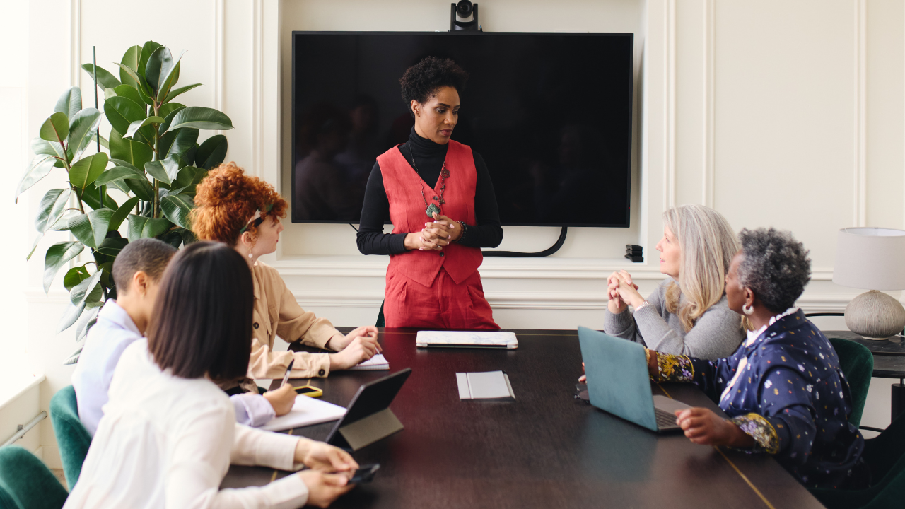 A photo of a Black woman at the head of a conference room table leading a meeting including five diverse individuals. This photo supports an article highlighting the importance of clearly communicating your goals as a leader in higher ed to get your team to buy into the long-term vision.