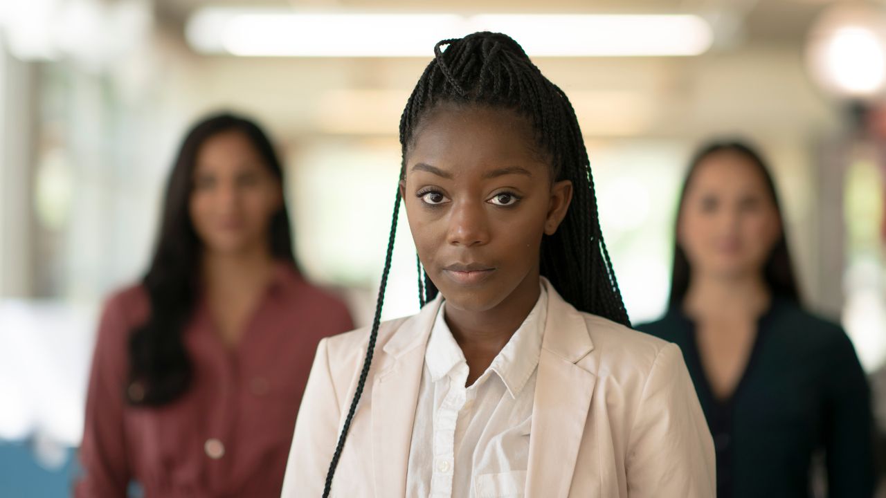black woman stands alone in front of two other women of color, staring at the camera, in an example of conflict management