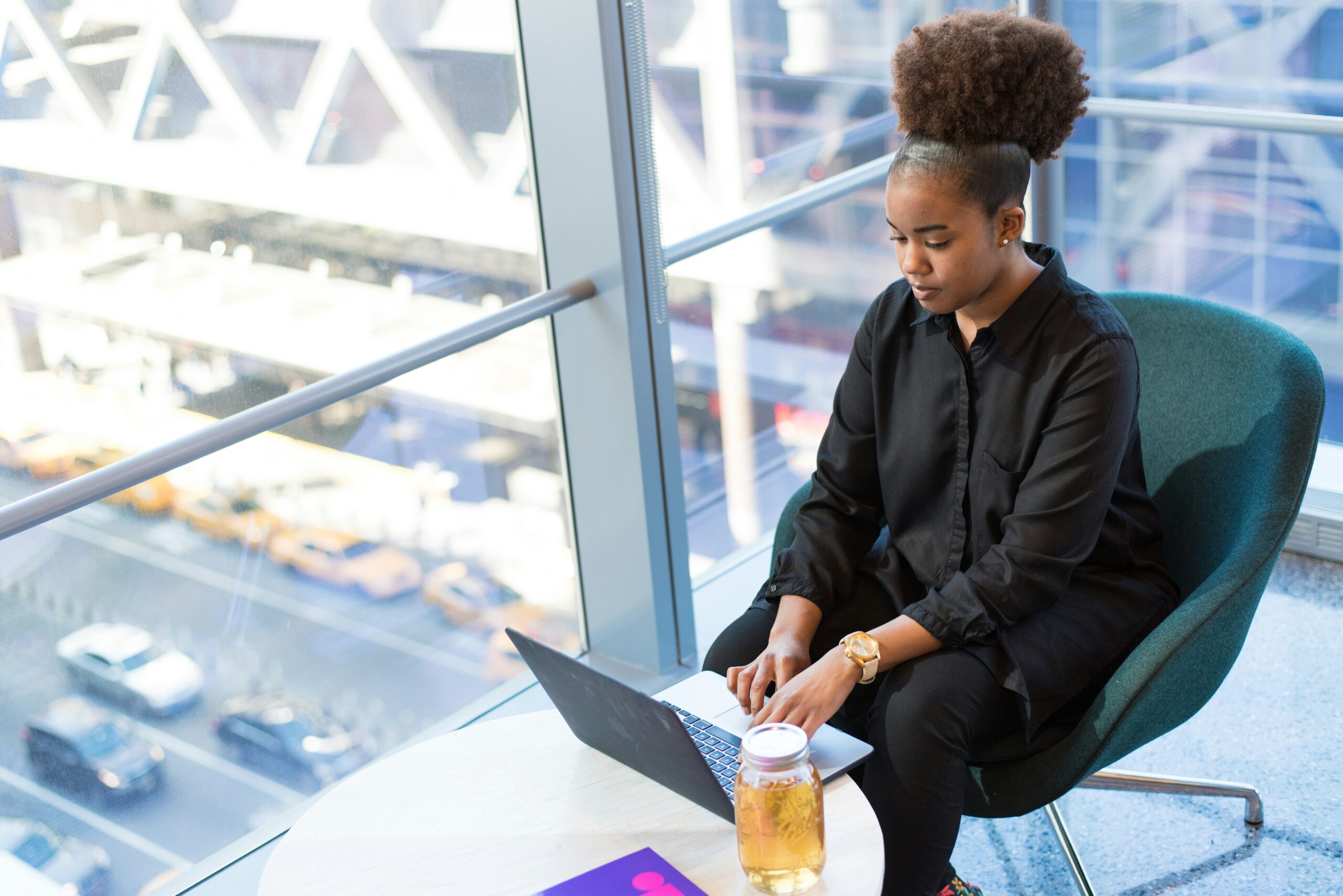 A woman of color sits on a chair with her laptop in front of her. She’s intently focused on the laptop. In the background, there is a big window showing a parking lot. Next to her, she has a glass of juice. This image accompanies a blog about reflecting on the past semester to improve leadership skills in women of color in higher education.